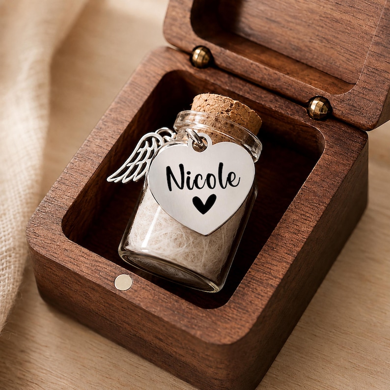May include: A small glass bottle with a cork stopper inside a dark wooden box. The bottle features a silver heart-shaped charm engraved with the name Nicole and a small heart symbol, along with a silver angel wing charm attached to the top.