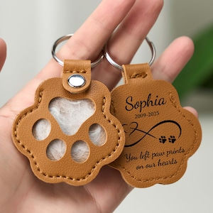 May include: Two tan leather paw print keychains. One keychain has a clear window, the other has the name "Sophia" and the years "2009-2025" with an infinity symbol and paw prints. The text reads "You left paw prints on our hearts."