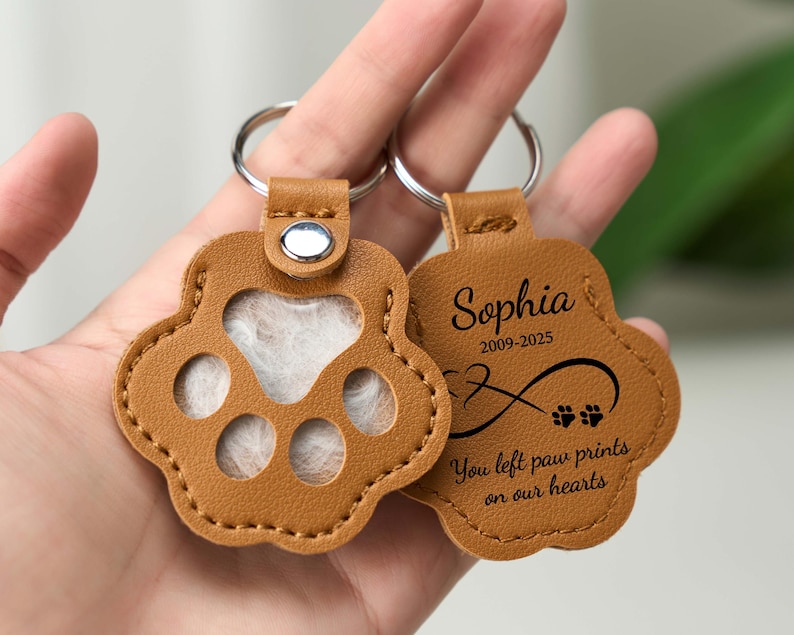 May include: Two tan leather paw-shaped keychains. One keychain has a clear window, the other has the name "Sophia", dates, an infinity symbol, and the text "You left paw prints on our hearts". Each keychain has a silver keyring.