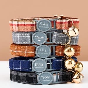 May include: Five cat collars in a stack, each with a different plaid pattern and colour: red, grey, brown, blue, and black. Each collar has a grey buckle with a name and phone number or instructions. Gold bells are attached.