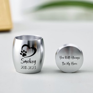 Op de afbeelding: Een zilveren urn voor huisdieren met een pootafdruk hart ontwerp en de naam "Smokey" met de jaren 2011-2023. Het deksel heeft de inscriptie "You Will Always Be My Hero."