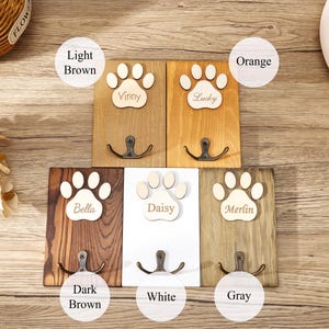 May include: Five rectangular wooden wall hooks featuring a paw print cutout at the top. Each hook is labeled with a name: Vinny, Lucky, Bella, Daisy, and Merlin. The wood finishes include light brown, orange, dark brown, white, and gray colors.