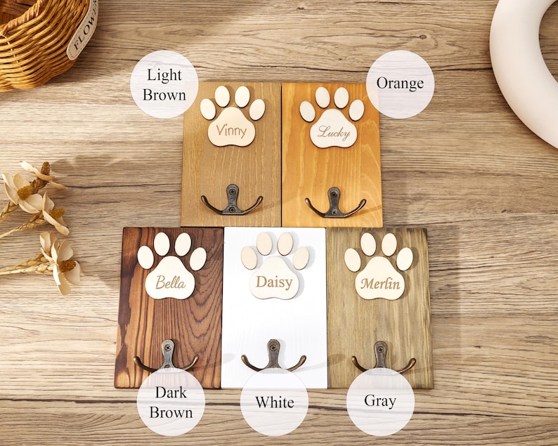 May include: Five rectangular wooden wall hooks featuring a paw print cutout at the top. Each hook is labeled with a name: Vinny, Lucky, Bella, Daisy, and Merlin. The wood finishes include light brown, orange, dark brown, white, and gray colors.