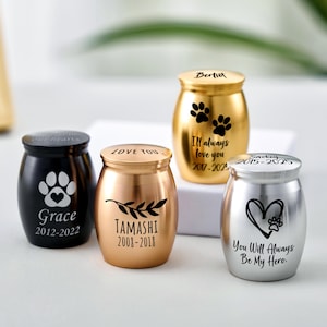 May include: Four small pet urns in gold, silver, and black. Each urn features unique designs, including paw prints, hearts, and inscriptions like "Grace 2012-2022" and "You Will Always Be My Hero."