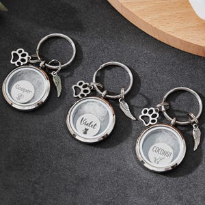 May include: Three silver keychains with round, clear lockets. Each keychain has a paw print charm, a wing charm, and a circular locket with a name inside: "Cooper", "Violet", and "Coconut". The keychains are on a dark surface.