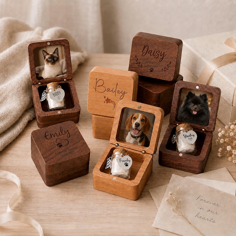 May include: A collection of square wooden keepsake boxes with hinged lids, some featuring engraved names like Daisy, Bailey, and Emily. Inside, small glass jars with cork stoppers and heart-shaped tags are displayed alongside pet photos.