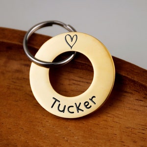 Pode incluir: Uma placa de identificação para cães de metal dourado com um coração e o nome "Tucker" gravado nela. A placa está presa a um chaveiro prateado.