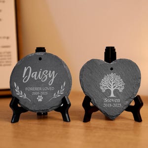 May include: Two slate memorial stones, one round and one heart-shaped, on black stands. The round stone is engraved with "Daisy" and "Forever Loved," with a paw print. The heart-shaped stone features a tree design and the name "Steven."