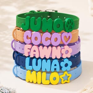 May include: A stack of five silicone pet collars in green, orange, purple, dark blue, and light blue. Each collar features large, raised, colourful letters spelling the names JUNO, COCO, FAWN, LUNA, and MILO, accompanied by paw, heart, flower, and star icons.
