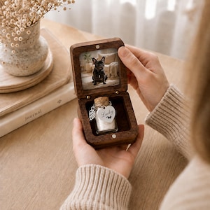 May include: A square dark wood box with a hinged lid featuring a photograph of a French Bulldog inside. The base contains a small glass bottle with a cork stopper, a silver angel wing charm, and a heart-shaped tag engraved with Lucky.