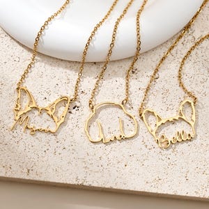Pode incluir: Três colares dourados com pingentes em forma de cão e correntes delicadas. Cada pingente apresenta o contorno de uma raça de cão diferente com um nome inscrito: "Megan", "Linda" e "Bella". Os colares são exibidos em uma superfície de tom neutro.