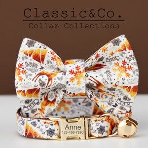 Puede incluir: Un collar de gato blanco con una hebilla dorada y una campanilla. El collar tiene una pajarita a juego con un fondo blanco y un patr&oacute;n de copos de nieve, ciervos y estrellas en naranja, rojo y gris. El collar est&aacute; personalizado con el nombre "Anne" y un n&uacute;mero de tel&eacute;fono.