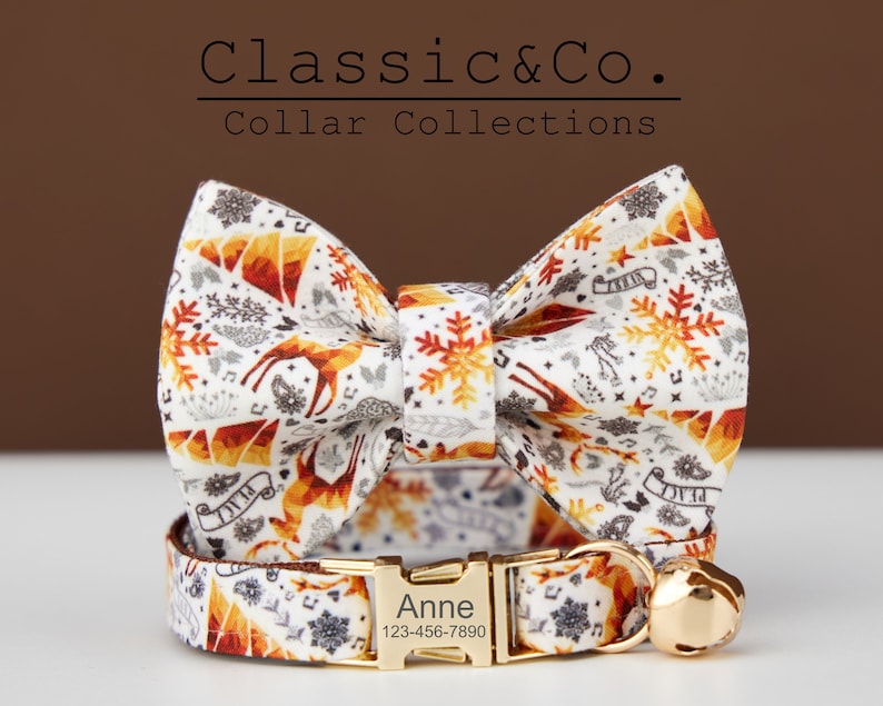 Puede incluir: Un collar de gato blanco con una hebilla dorada y una campanilla. El collar tiene una pajarita a juego con un fondo blanco y un patr&oacute;n de copos de nieve, ciervos y estrellas en naranja, rojo y gris. El collar est&aacute; personalizado con el nombre "Anne" y un n&uacute;mero de tel&eacute;fono.