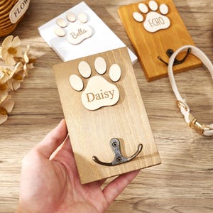May include: A rectangular wooden wall hook featuring a light-colored wooden paw print cutout with the name Daisy engraved in the center. A dark metal double hook is attached to the lower portion of the wood plaque, which has a natural wood grain finish.