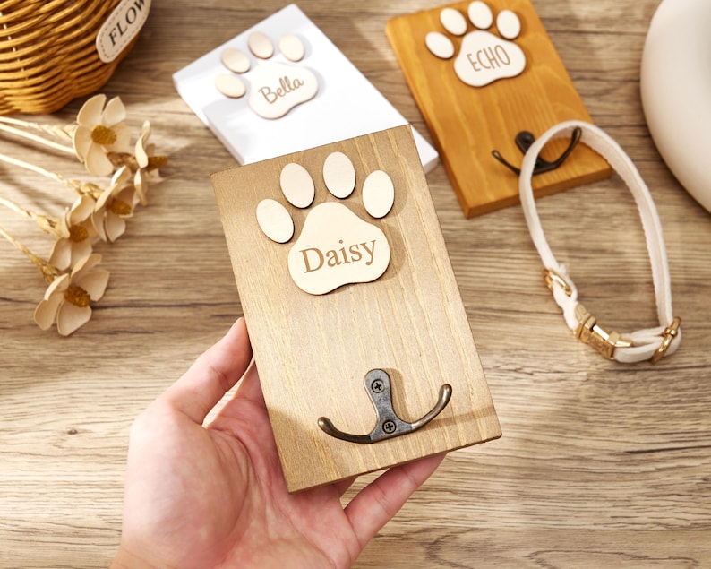 May include: A rectangular wooden wall hook featuring a light-colored wooden paw print cutout with the name Daisy engraved in the center. A dark metal double hook is attached to the lower portion of the wood plaque, which has a natural wood grain finish.
