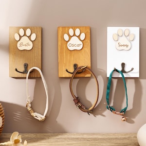 May include: Three rectangular wall-mounted wooden plaques, each featuring a light-colored wooden paw print cutout with the engraved names Busta, Oscar, and Sammy. Each plaque includes a dark metal double hook holding a pet collar in white, brown, or green.
