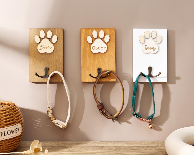 May include: Three rectangular wall-mounted wooden plaques, each featuring a light-colored wooden paw print cutout with the engraved names Busta, Oscar, and Sammy. Each plaque includes a dark metal double hook holding a pet collar in white, brown, or green.
