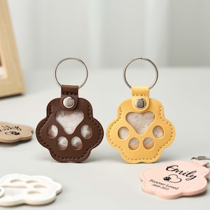 May include: Two paw-shaped keychains, one brown and one yellow, with silver keyrings. The paw pads are white. A light pink keychain with the name "Emily" and the text "Forever Loved" is also visible.