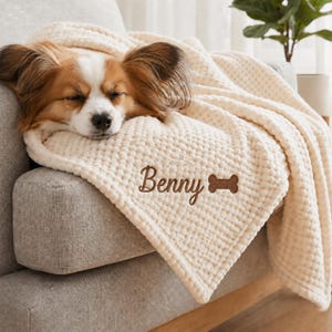 May include: A cream-colored waffle-textured blanket featuring the embroidered name Benny in brown script lettering next to a brown embroidered dog bone icon. A small dog with brown and white fur is resting its head on the soft fabric.