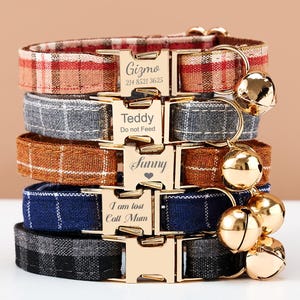 May include: A stack of personalized cat collars in various plaid patterns. Each collar has a gold-colored buckle with engraved text, including names and phone numbers. Gold bells hang from the collars.