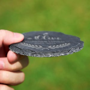 May include: A round, dark grey slate coaster with a textured edge. The coaster features etched text and a laurel wreath design. The coaster is held in a hand against a blurred green background.