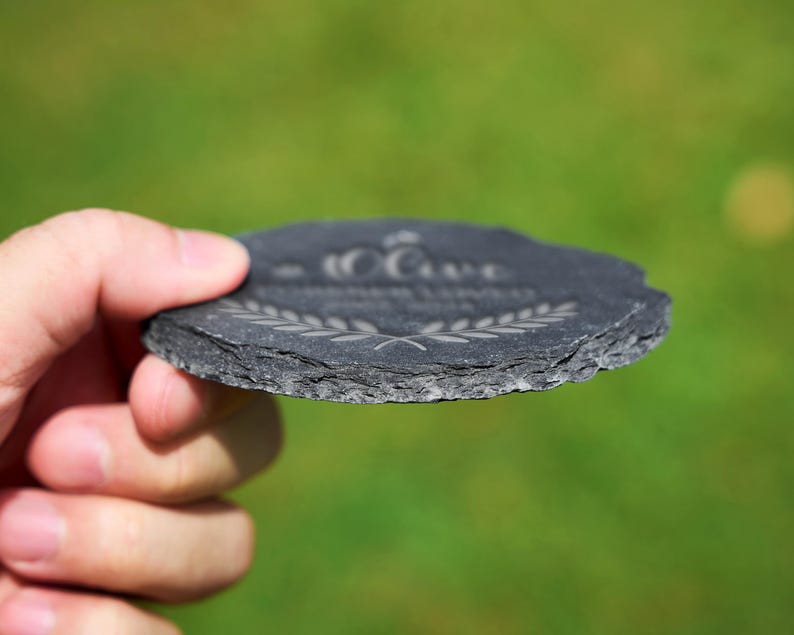 May include: A round, dark grey slate coaster with a textured edge. The coaster features etched text and a laurel wreath design. The coaster is held in a hand against a blurred green background.