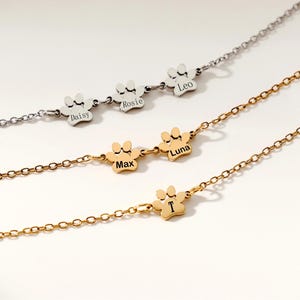 May include: Three personalized paw print necklaces in silver and gold tones. Each necklace features a chain and paw print charm with a name engraved on it. The names Daisy, Rosie, Leo, Max, Luna, and T are visible.