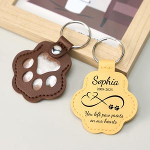 May include: Two paw-shaped keychains. One is brown leather with a clear window, the other is yellow leather with the name "Sophia", dates, an infinity symbol, and the text "You left paw prints on our hearts". Both have silver key rings.