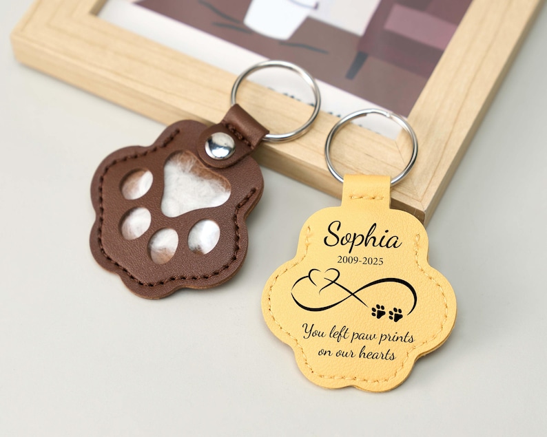May include: Two paw-shaped keychains. One is brown leather with a clear window, the other is yellow leather with the name "Sophia", dates, an infinity symbol, and the text "You left paw prints on our hearts". Both have silver key rings.