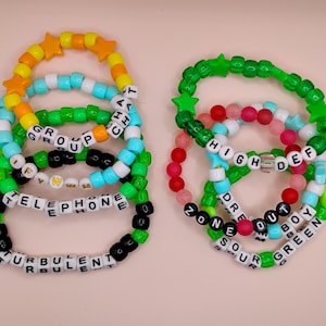 May include: A collection of colorful beaded bracelets. The bracelets feature various colors including green, orange, blue, and black. Some bracelets have star-shaped beads and text that reads "GROUP," "TELEPHONE," "TURBULENT," "HIGH DEF," "DREAM BOY," and "SOUR GREEN."