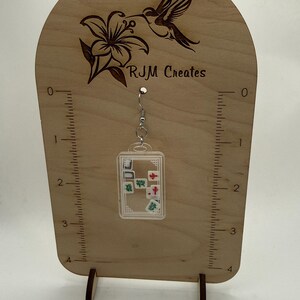 May include: Clear acrylic earrings with miniature mahjong tiles inside, hanging from a silver-colored hook. The earrings are displayed on a wooden stand with a hummingbird and flower design, and the text "RJM Creates".