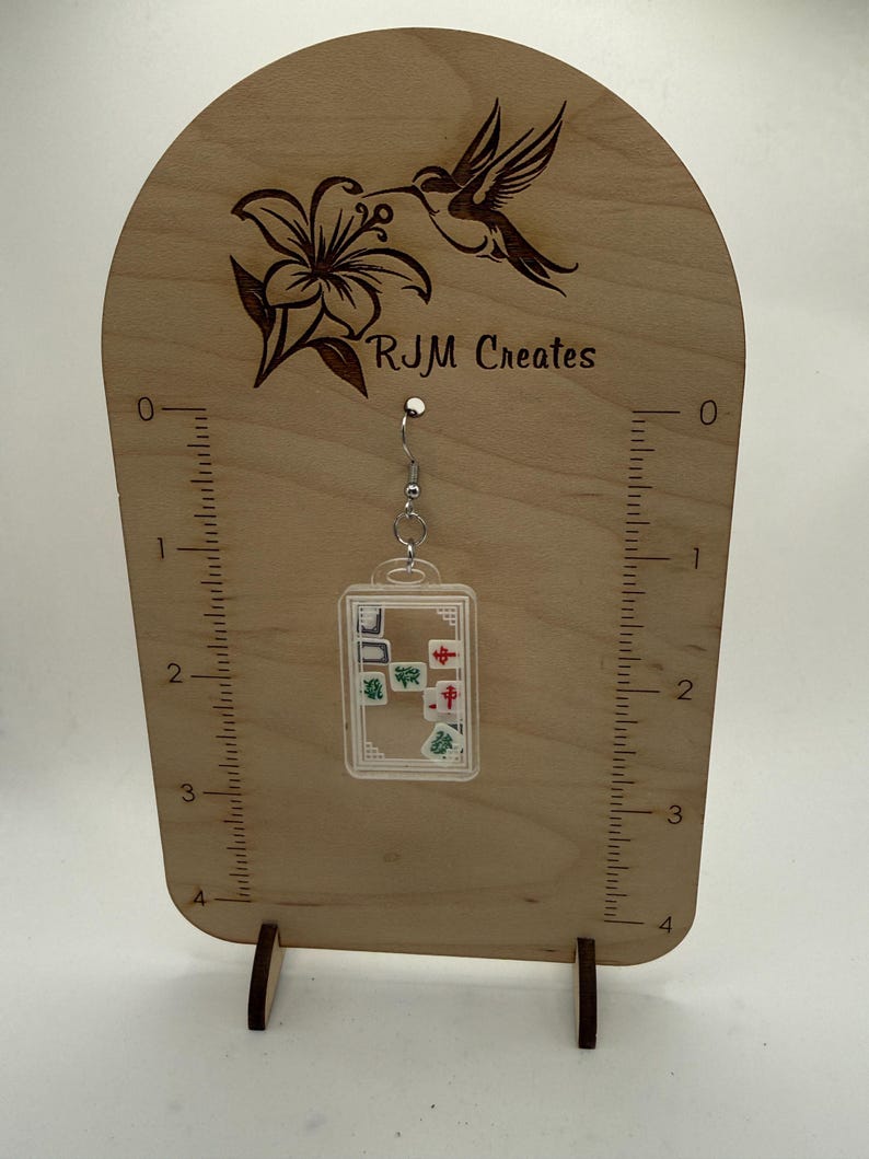 May include: Clear acrylic earrings with miniature mahjong tiles inside, hanging from a silver-colored hook. The earrings are displayed on a wooden stand with a hummingbird and flower design, and the text "RJM Creates".