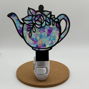May include: A decorative night light featuring a teapot design. The teapot is black with a stained glass-like pattern in shades of blue, purple, and pink. It is mounted on a white plug-in base and a round cork base.