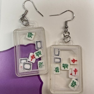 May include: Clear rectangular earrings with silver hooks, each containing miniature Mahjong tiles. The tiles feature red, green, and black symbols. The earrings are displayed on a white and purple background.