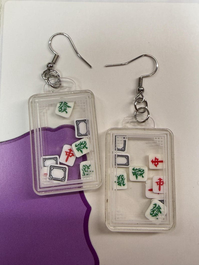 May include: Clear rectangular earrings with silver hooks, each containing miniature Mahjong tiles. The tiles feature red, green, and black symbols. The earrings are displayed on a white and purple background.