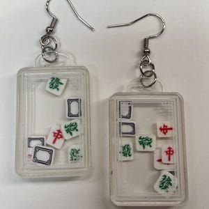 May include: A pair of clear rectangular earrings with silver hooks. Each earring contains miniature Mahjong tiles in various colors, including green, red, and black, within a clear frame. The tiles are scattered inside the frame.