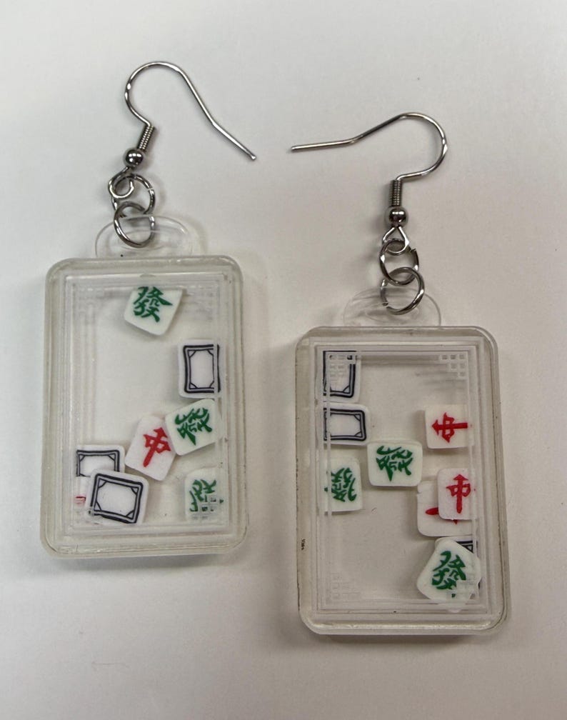 May include: A pair of clear rectangular earrings with silver hooks. Each earring contains miniature Mahjong tiles in various colors, including green, red, and black, within a clear frame. The tiles are scattered inside the frame.