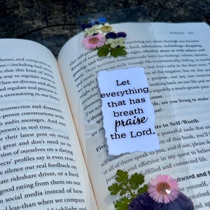 May include: Open book with a bookmark featuring dried flowers and the text "Let everything that has breath praise the Lord." The bookmark is made of clear plastic.
