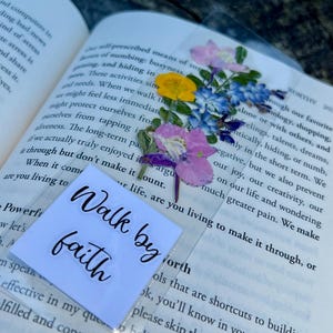 May include: A clear bookmark with dried flowers in shades of purple, yellow, and blue. The bookmark has the words "Walk by faith" printed on a white square. The bookmark is placed in an open book.
