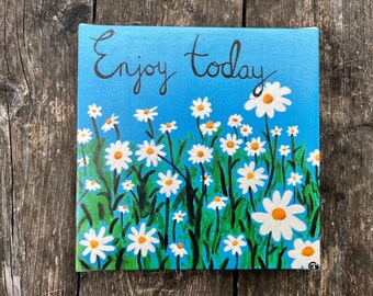 May include: A blue canvas painting with the text "Enjoy today" in black cursive lettering. The background is a light blue color with white daisies painted on top.