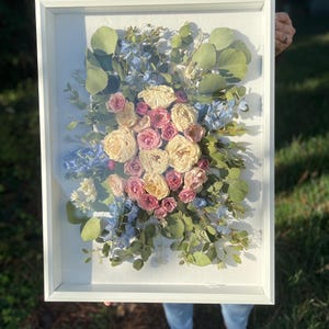 May include: A framed floral arrangement featuring a bouquet of dried flowers. The arrangement includes cream, pink, and blue flowers, with green eucalyptus leaves. The white frame enhances the colors of the flowers, creating a decorative piece.