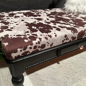 May include: A black wooden ottoman with a brown and white cowhide patterned top. The ottoman has a drawer with a silver knob and turned legs.