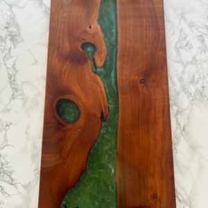 May include: A rectangular wooden serving tray with a dark brown finish and a central river of green resin. The tray has black metal handles on each end. The wood grain is visible, adding a natural aesthetic to the design.