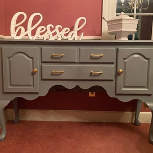May include: A gray painted buffet with gold hardware, featuring two doors and two drawers. The word "blessed" is displayed on top. A white serving dish with a lid sits on the right side of the buffet.