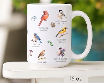 May include: White ceramic mug featuring illustrations of various birds, including a Northern Cardinal, Eastern Bluebird, and Ruby-Throated Hummingbird. The mug also has the text "15 oz option".