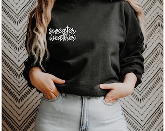May include: A black sweatshirt with the text "sweater weather" in white script on the front. The model is wearing the sweatshirt with blue jeans.