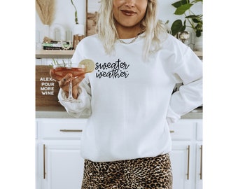 May include: A white sweatshirt with the text "sweater weather" in black script. The model is wearing the sweatshirt with a brown hat and leopard print pants.
