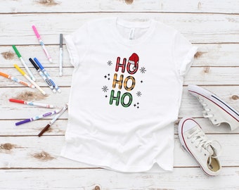 May include: White t-shirt featuring a red, green and yellow plaid "HO HO HO" design with a Santa hat on top of the first "HO".