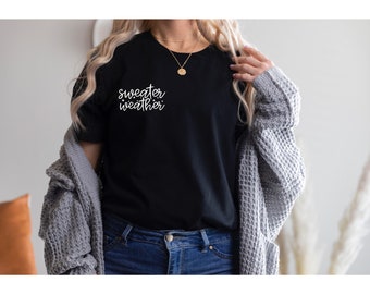 May include: A black t-shirt with the text "sweater weather" in white lettering. The shirt is being worn by a person with a gray cardigan and blue jeans.