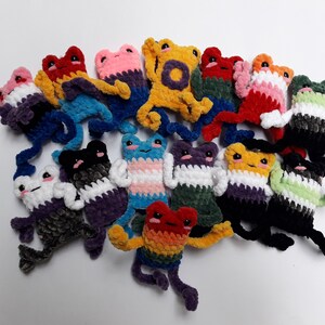 Peut inclure: Une collection de peluches de grenouilles au crochet dans différentes couleurs et motifs, notamment arc-en-ciel, fierté de genre et noir et blanc. Les grenouilles sont disposées en cercle sur un fond blanc.